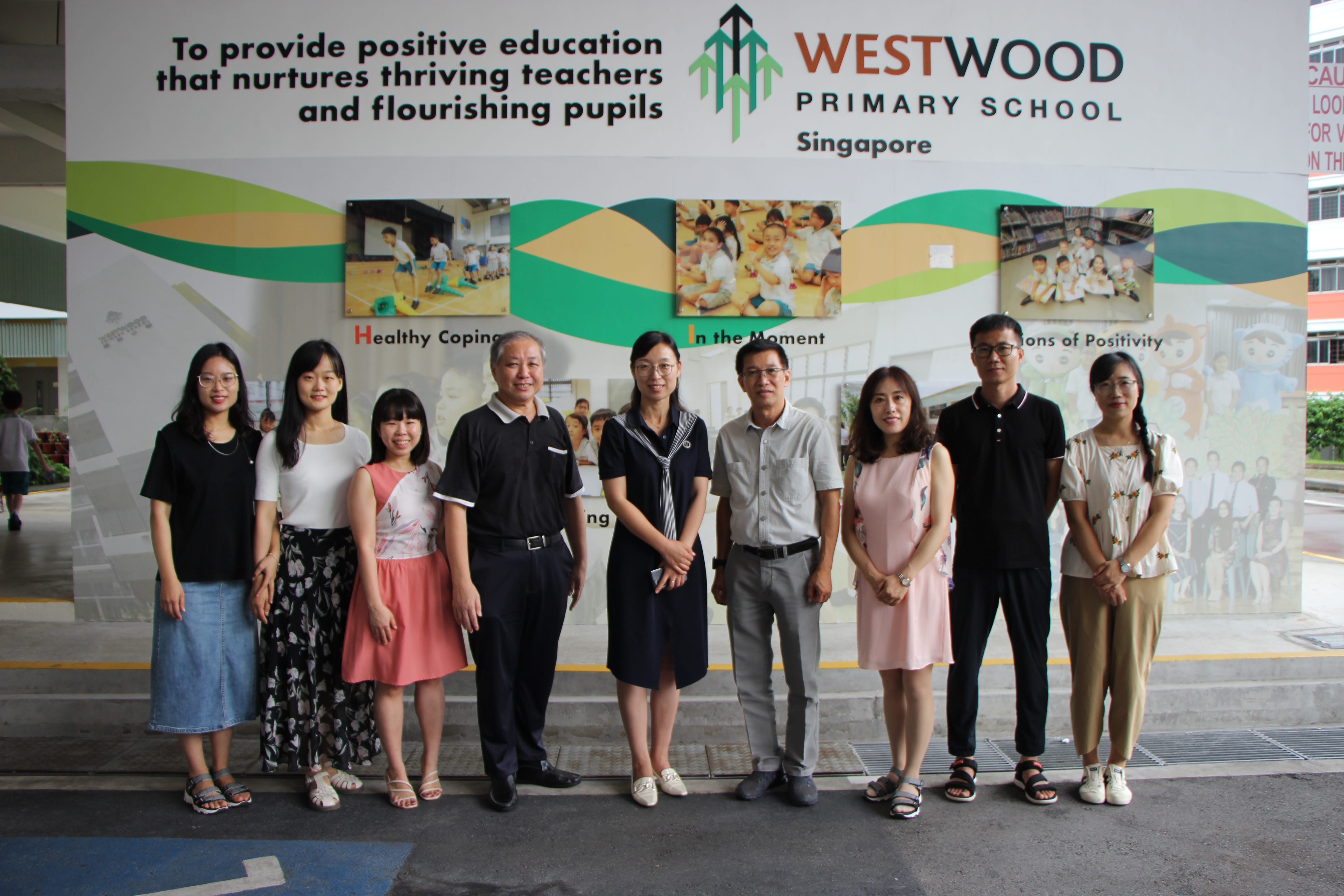 Eight adults pose together in front of a Westwood Primary School Singapore wall display.